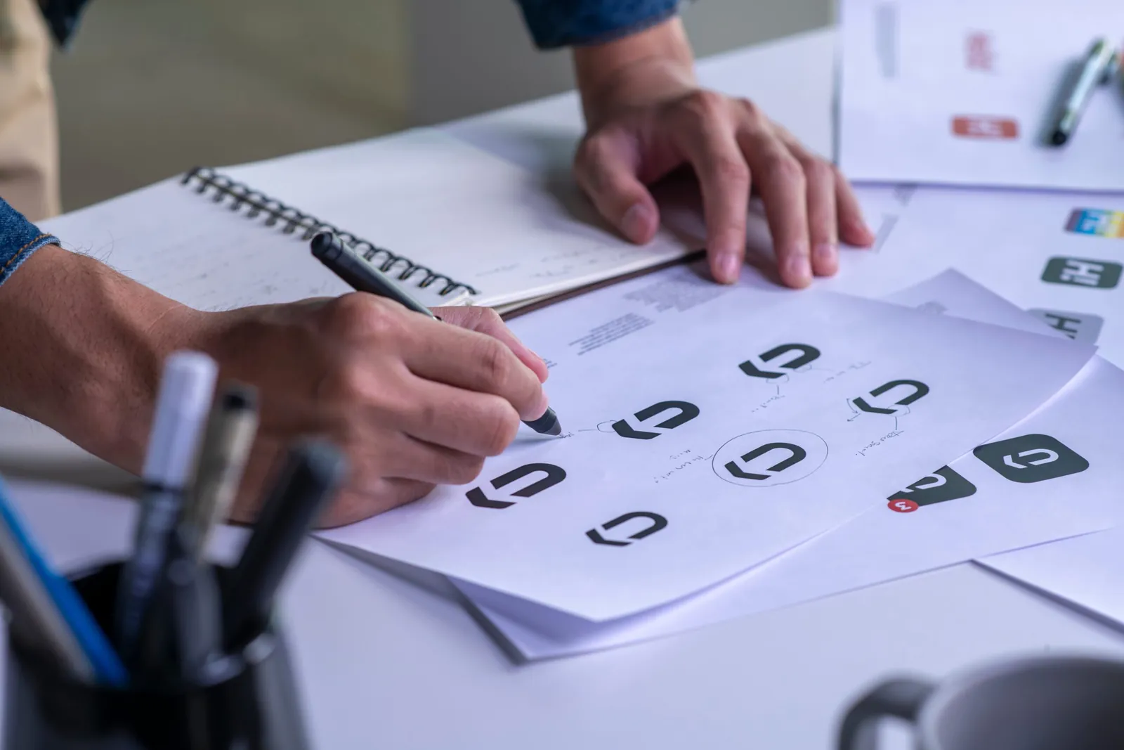 Tervező filctollal logóvázlatokat rajzol, mellette jegyzetfüzet és nyomtatott minták